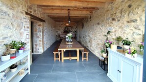 Un couloir aux murs de pierre mène à un espace repas où se trouvent une table et des bancs en bois, de part et d’autre desquels sont disposées des étagères ornées de plantes en pot.