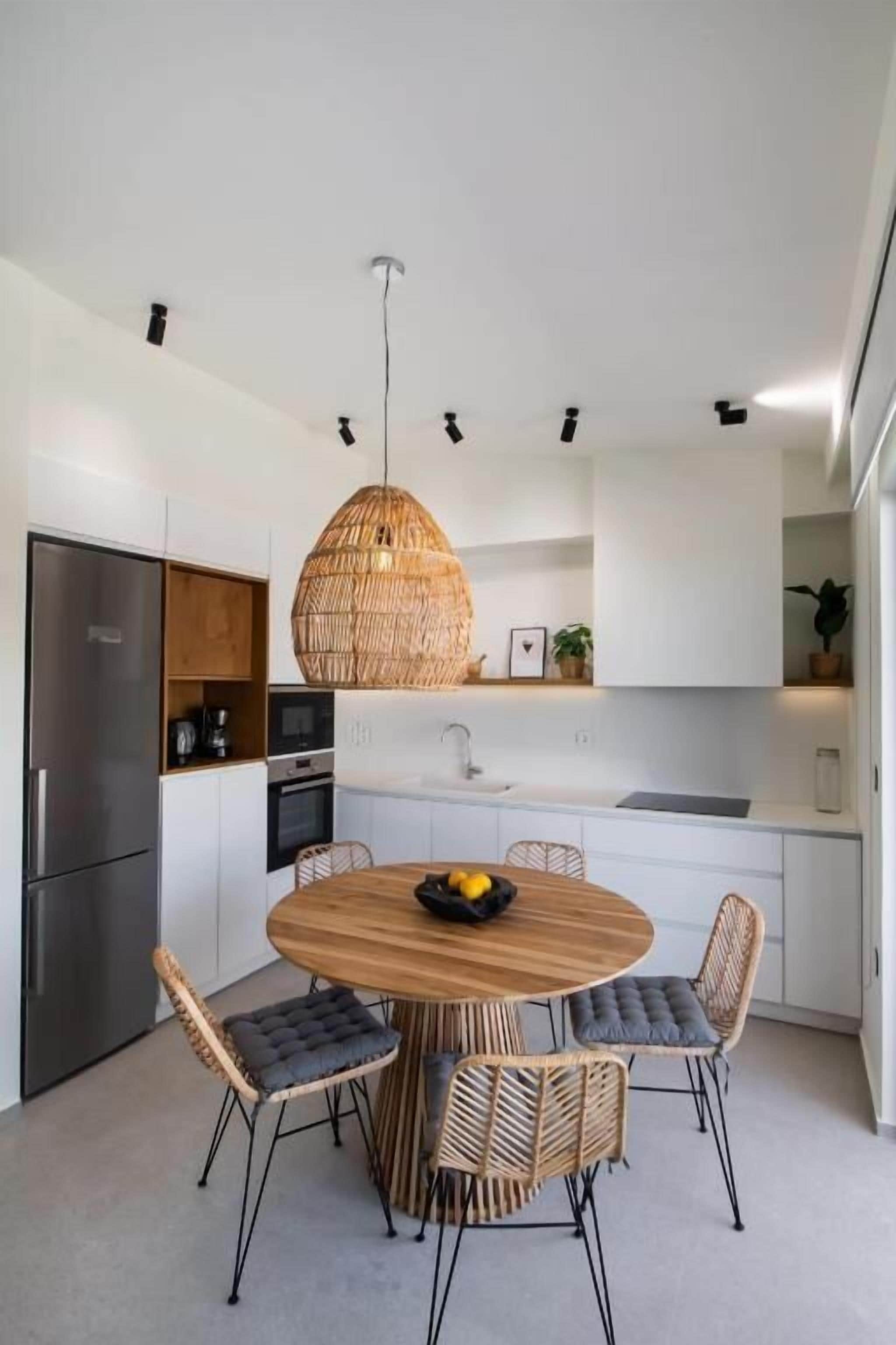 A modern kitchen with a round wooden table, wicker chairs, and a hanging light fixture.