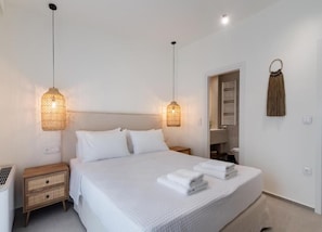 Un llit ben arropat amb llençols blancs i tovalloles doblegades, dues làmpades de nit, una tauleta de nit de fusta i una decoració de borrons penjada a la paret.