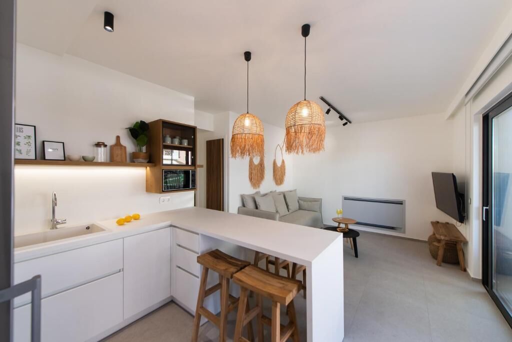 Una cocina moderna con armarios blancos, una isla central con taburetes, luces colgantes y una zona de comedor con sofá y televisor.