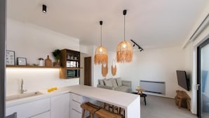 Una cocina moderna con armarios blancos, una isla central con taburetes, luces colgantes y una zona de comedor con sofá y televisor.