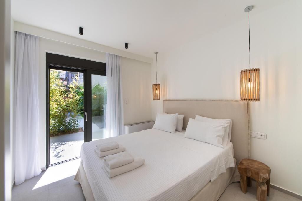 Ein Schlafzimmer mit einem großen Bett, einem hölzernen Hocker, einer Deckenleuchte und Blick durch eine Glastür.