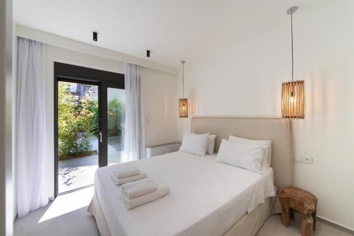 Une chambre avec un grand lit, un tabouret en bois, un luminaire au plafond et une vue à travers une porte vitrée.