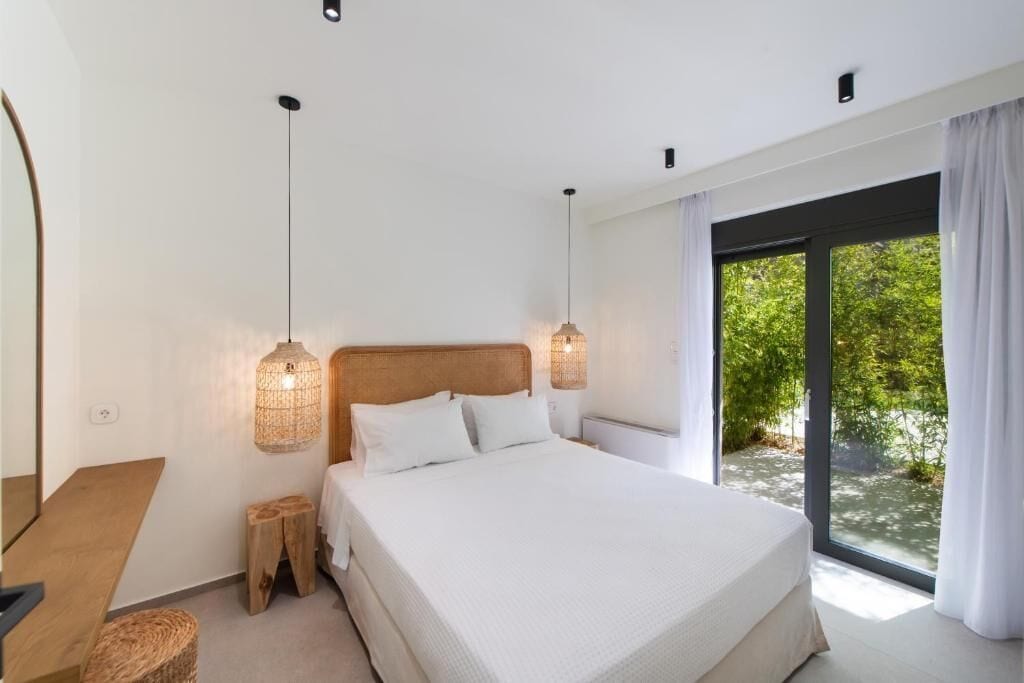 Un dormitorio con una cama grande, lámparas de noche, un cabecero de madera, un taburete pequeño y vistas a zonas verdes a través de una puerta de cristal.