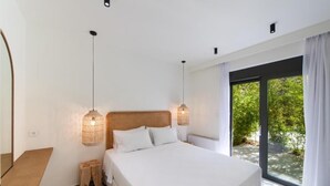 Un dormitorio con una cama grande, lámparas de noche, un cabecero de madera, un taburete pequeño y vistas a zonas verdes a través de una puerta de cristal.