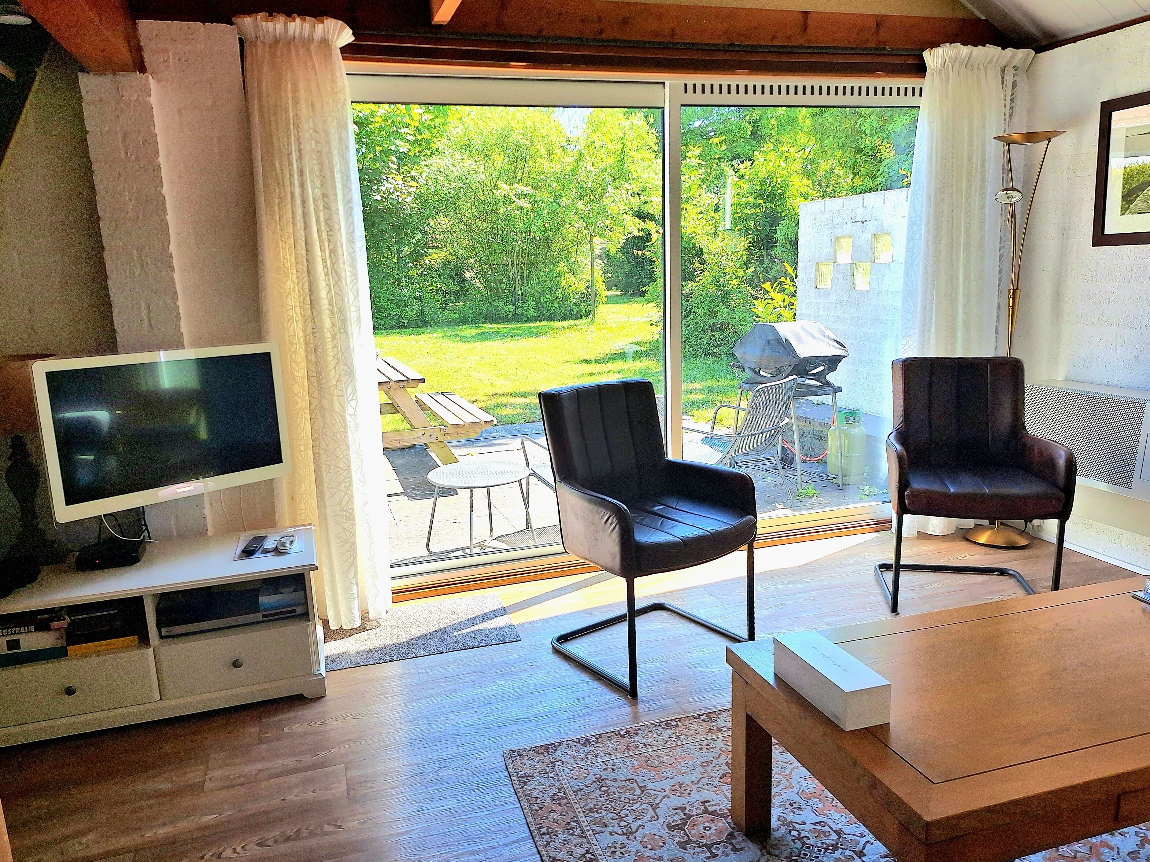 A living room with a TV, two chairs, a table, and a view of a garden through a sliding door.