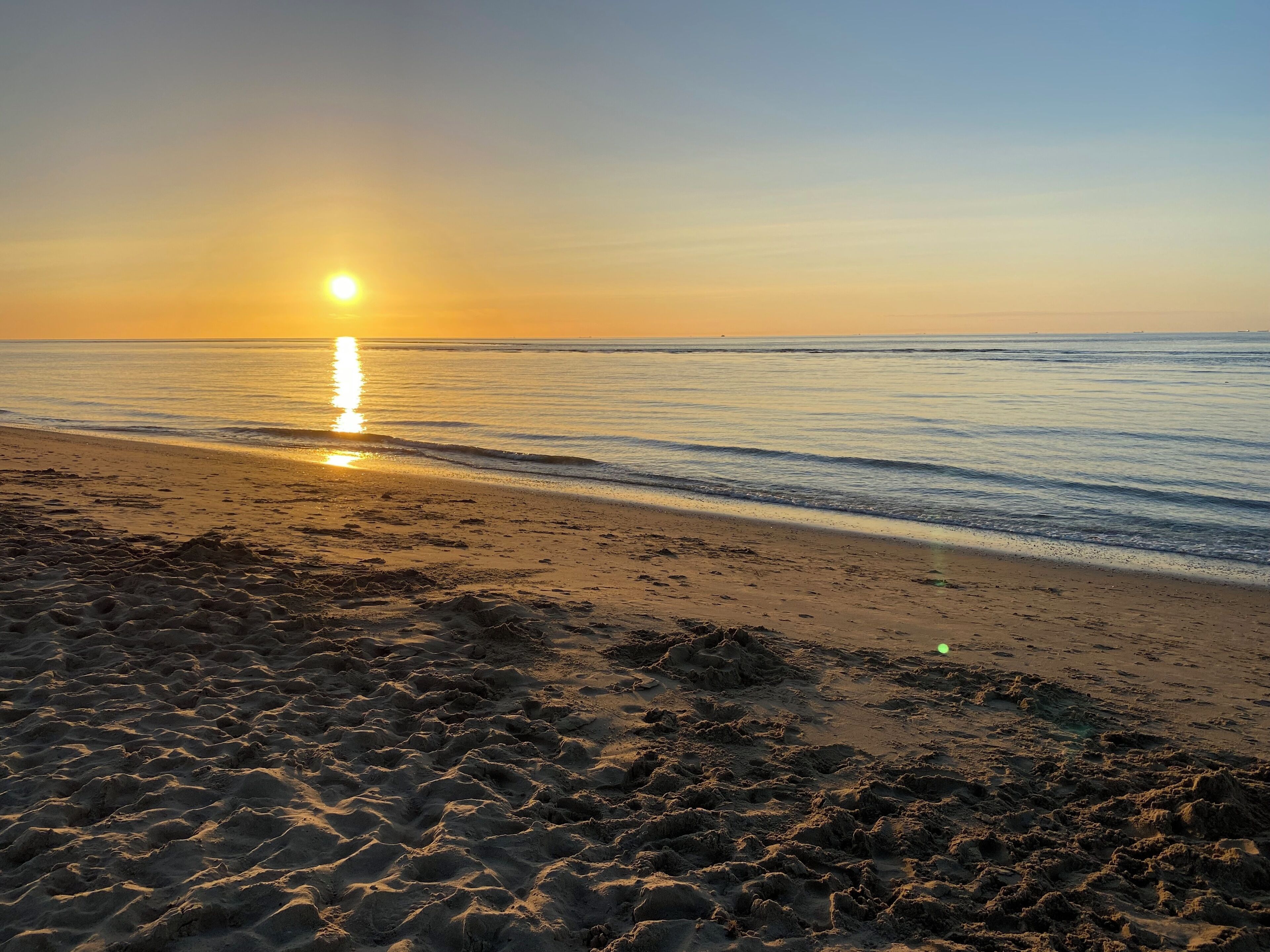 A beach with the sun setting over the horizon, calm sea, and footprints in the sand.