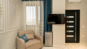 A living room with a beige sofa, a flat-screen TV, a small refrigerator, and a door with a handle. - Klegis Room (Durrës)