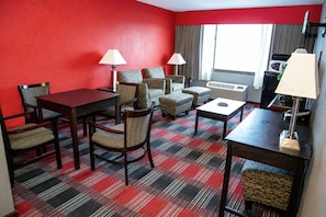 Habitación de hotel con pared roja, alfombra a cuadros, mesa de comedor con sillas, un conjunto de sofás, dos lámparas y una ventana con cortinas.