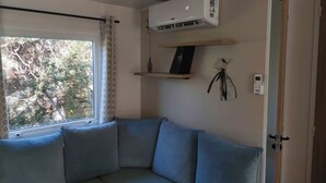 A room with a corner sofa, a window with curtains, an air conditioning unit, a wall-mounted light, and a door. - Rent a new mobile home at the 4-star Les Charmette campground—fully equipped (Les Mathes)