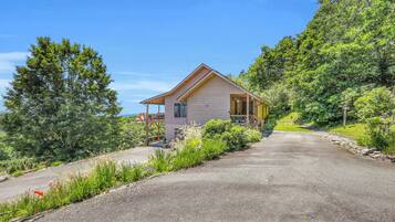 A single-story wooden cabin with a porch, surrounded by lush greenery and a clear sky.