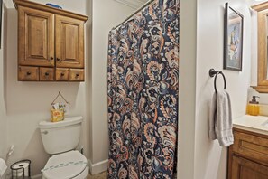 A bathroom with a toilet, wooden cabinets, a patterned shower curtain, a towel rack, and a framed picture on the wall.