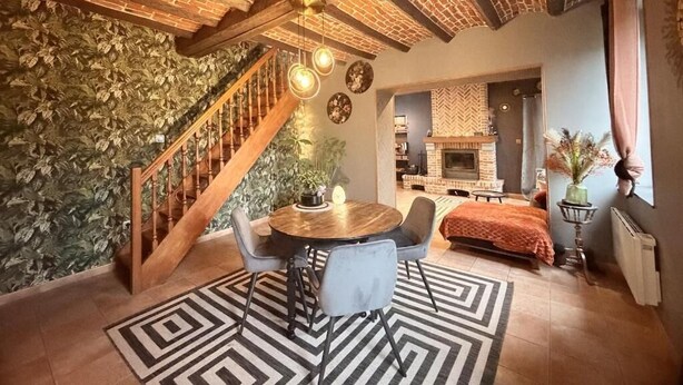 A cozy room with a wooden staircase, a round dining table with chairs, a fireplace, and a bed with an orange blanket. - La clos de la ch'tite d'alsace (Crespin)