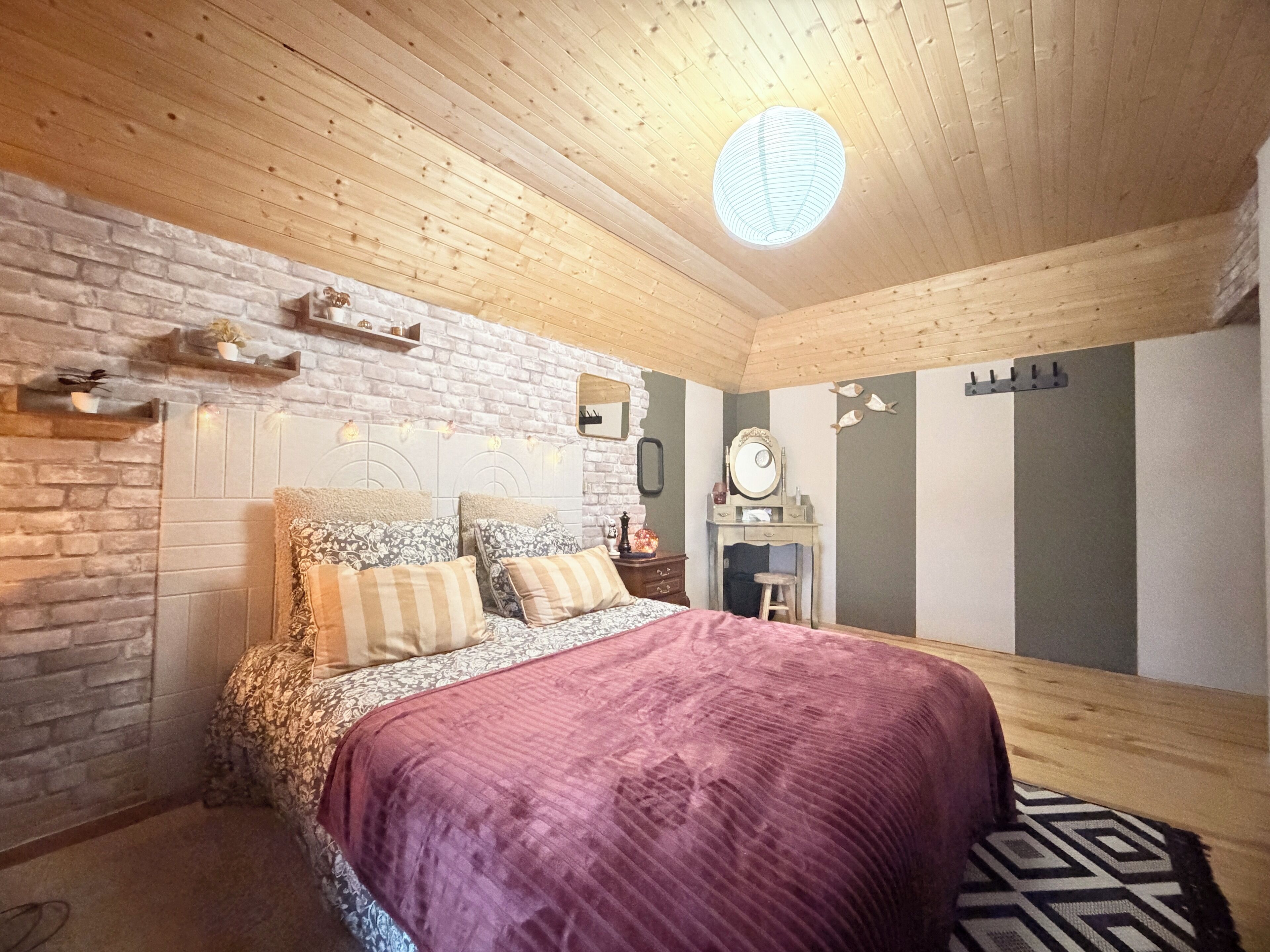 Quarto com teto de madeira, parede de tijolo, cama com roupa de cama estampada, mesa de cabeceira, cômoda e espelho.