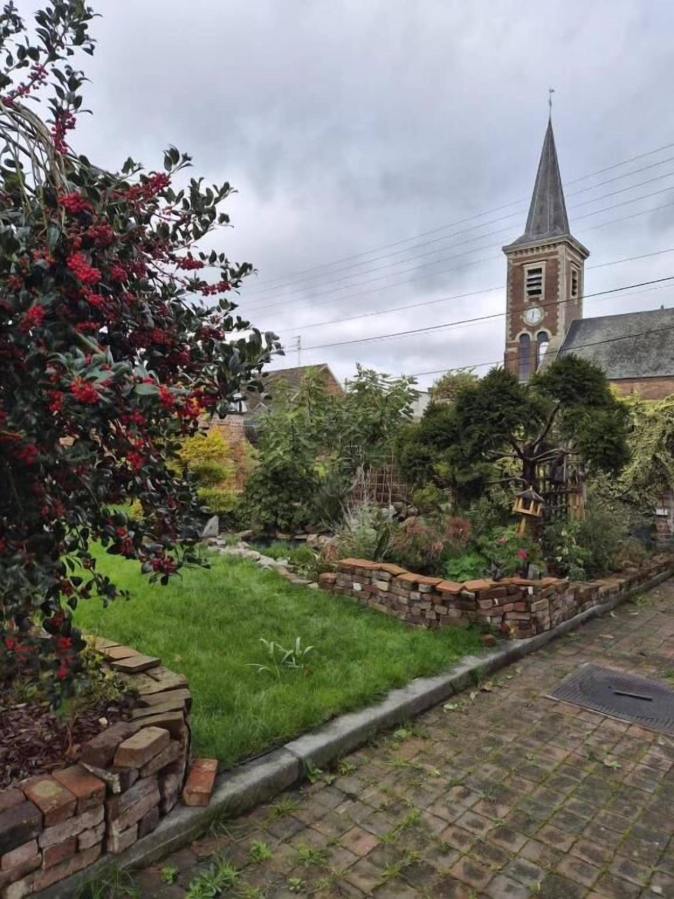 A church with a tall steeple, a brick wall, and a grassy area with trees.
