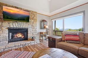 Ein Kamin aus Stein mit Feuer, ein großes Fernsehgerät darüber montiert, eine Couch mit einem gemusterten Kissen und ein Blick ins Freie durch große Fenster.