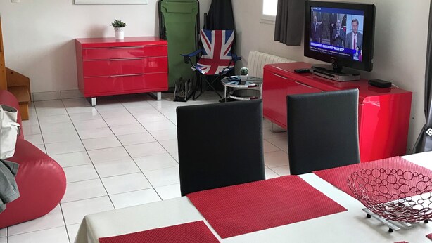 Uma sala de estar moderna com um sofá vermelho, uma mesa de jantar com jogos americanos vermelhos, uma televisão e uma cadeira verde dobrável.