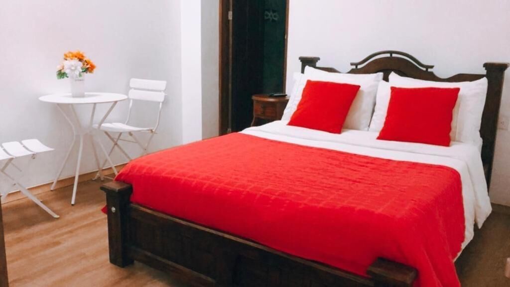 Un dormitorio con cama de madera, ropa de cama blanca y almohadas rojas. Hay una mesita blanca con una silla y un florero con flores.
