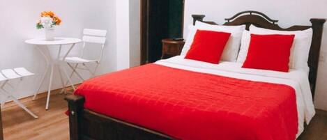Un dormitorio con cama de madera, ropa de cama blanca y almohadas rojas. Hay una mesita blanca con una silla y un florero con flores.
