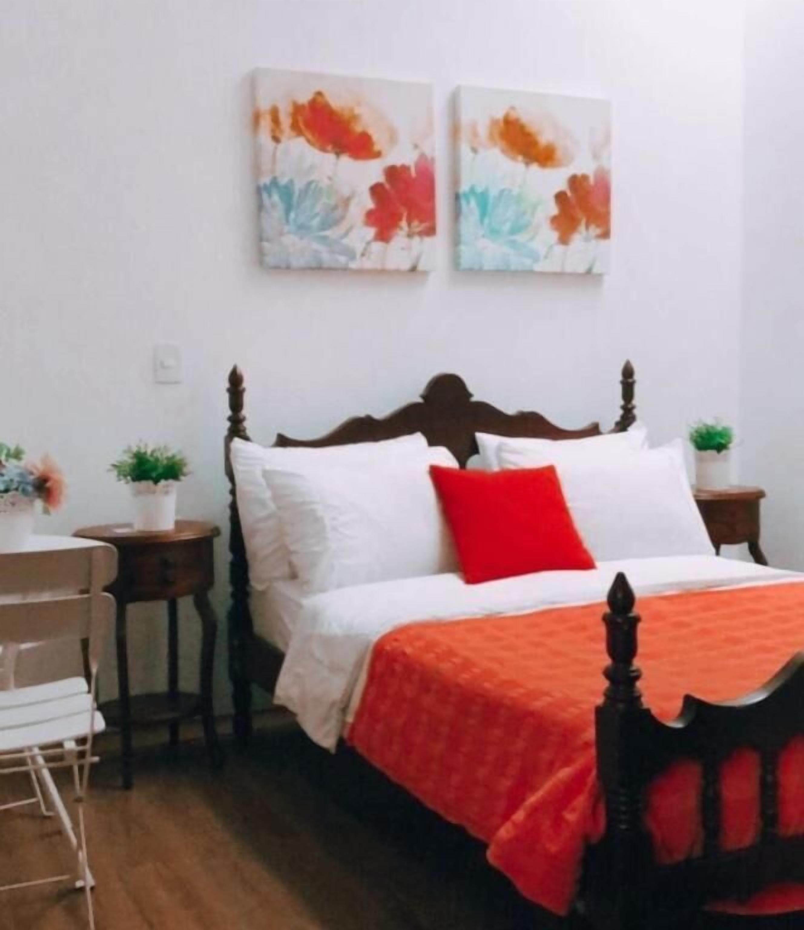 Slaapkamer met een houten bed, wit beddengoed, een rood kussen, twee aan de muur hangende schilderijen, een kleine plant en een stoel.