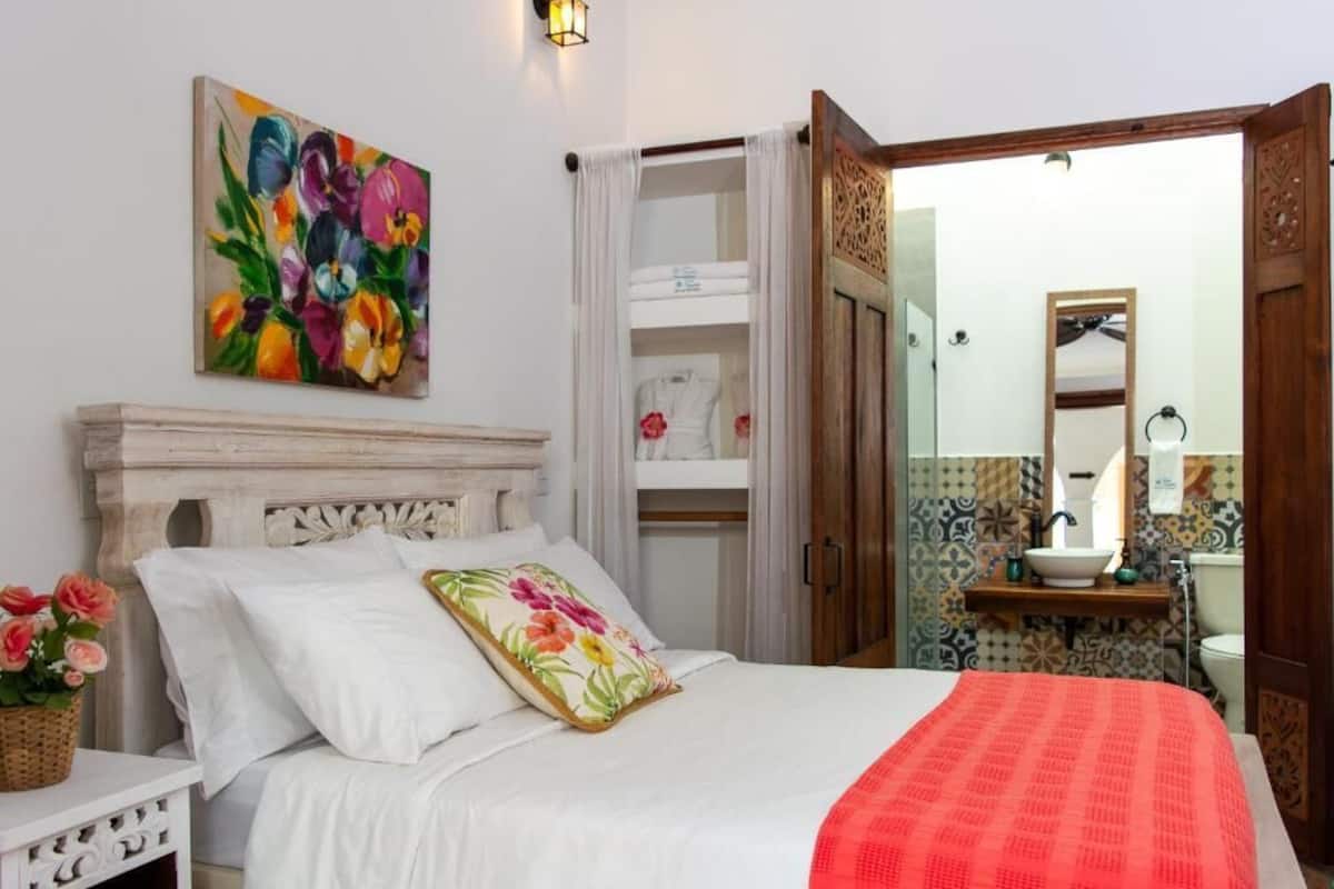 A bedroom with a bed, a painting, a nightstand, a bedside table, a mirror, and a bathroom visible through an open door.
