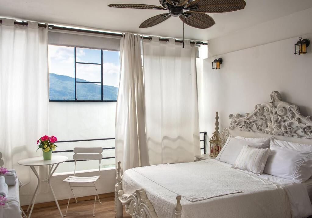 A bedroom with a bed, a window with curtains, a table with a chair, and a ceiling fan.