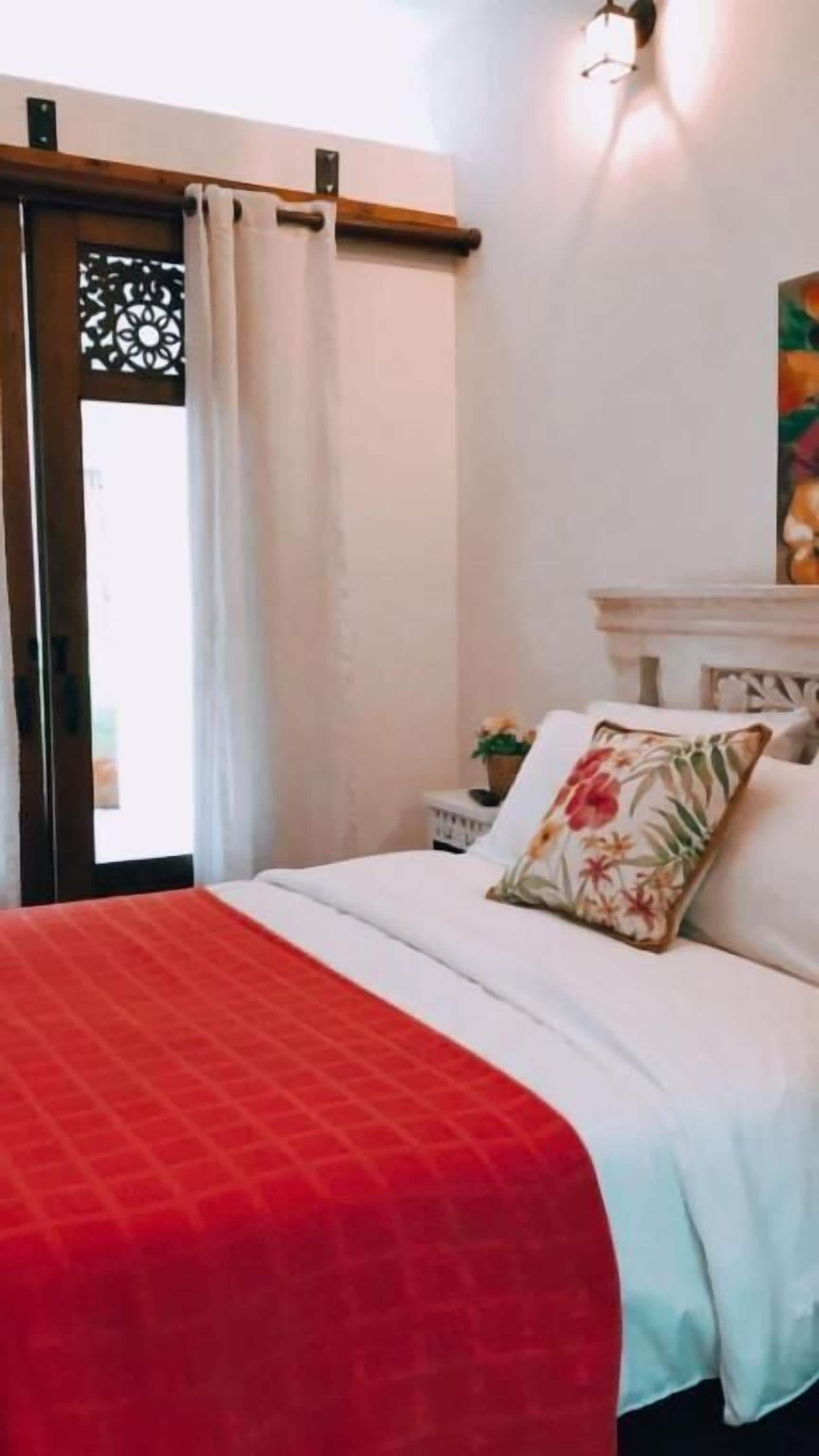 A bed with a red blanket, a white pillow with a floral design, and a nightstand with a plant.