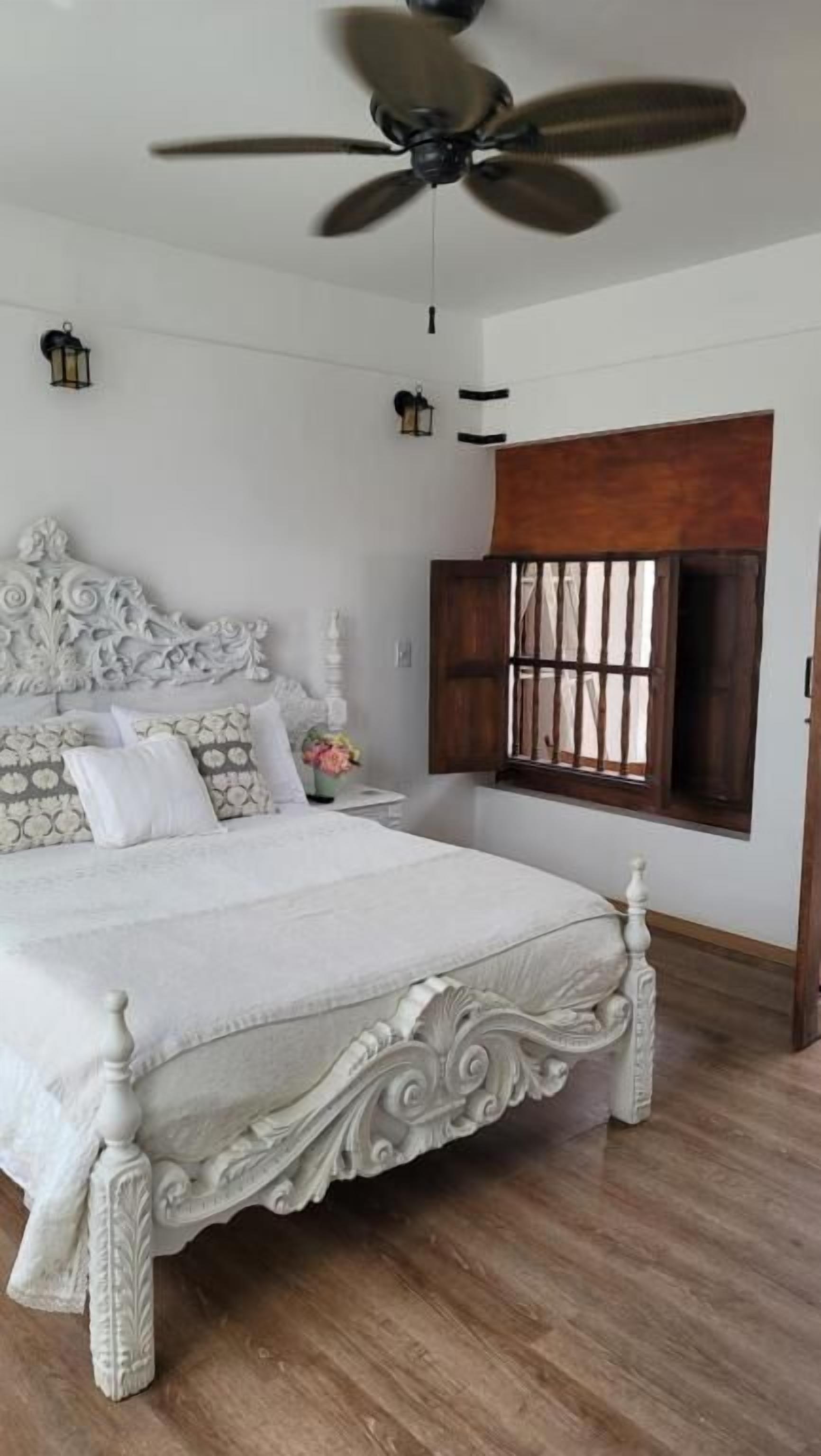 Un dormitorio con una cama grande, un ventilador de techo, una ventana con contraventanas de madera y una mesita de noche con un jarrón.