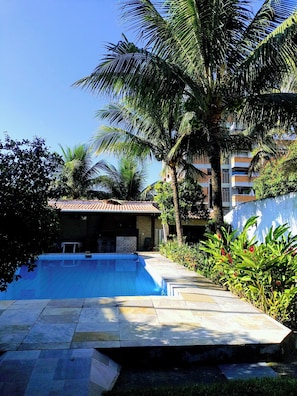 A swimming pool with a tiled deck, surrounded by palm trees and greenery, with buildings in the background.