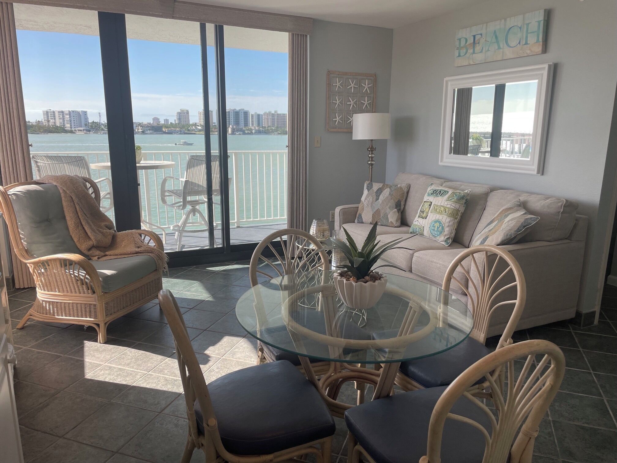 A room with a balcony, wicker furniture, a glass table, and a view of the city.