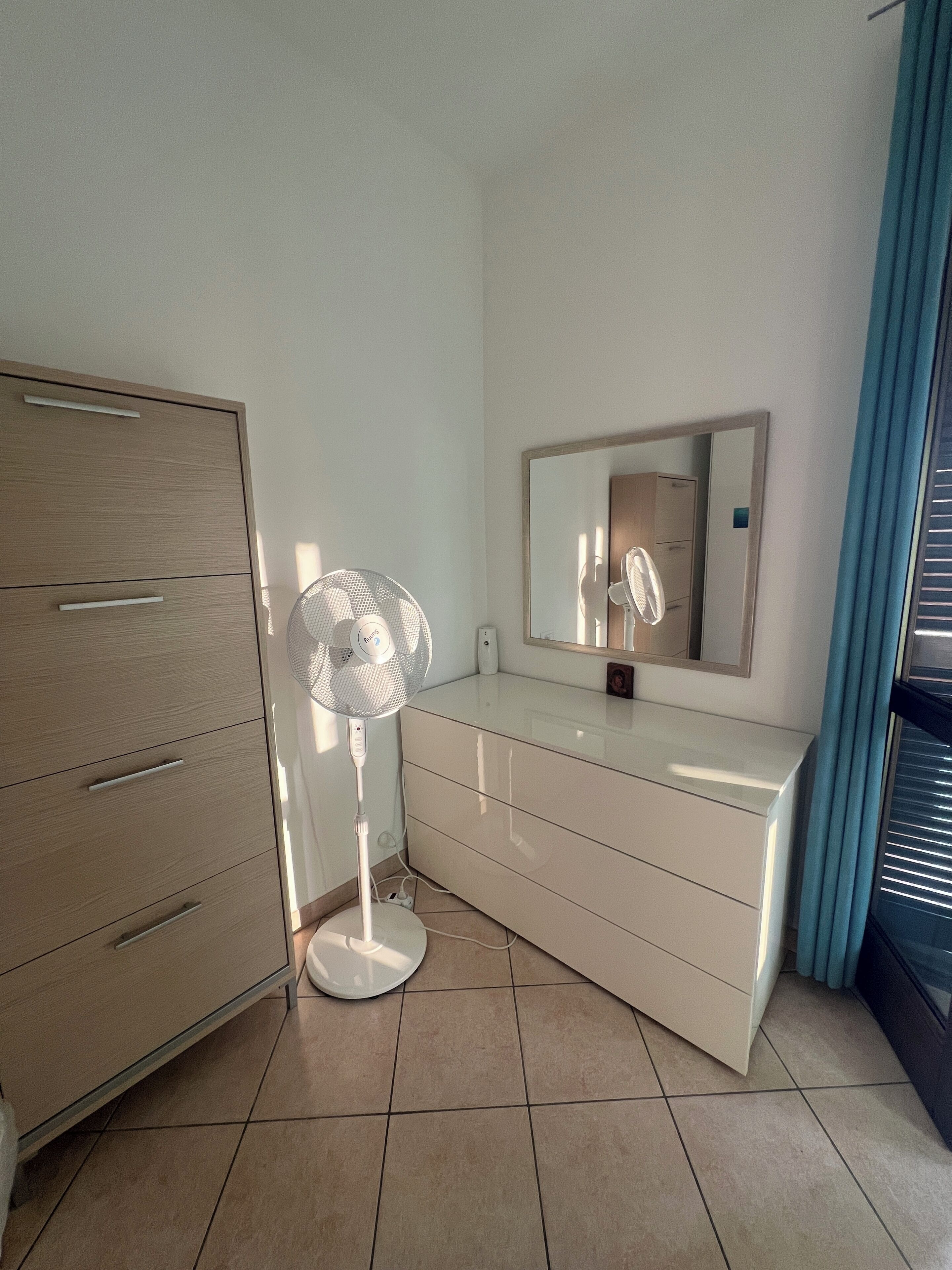 Une pièce comprenant une armoire en bois, une commode blanche, un ventilateur sur pied, un miroir et une fenêtre avec des stores.