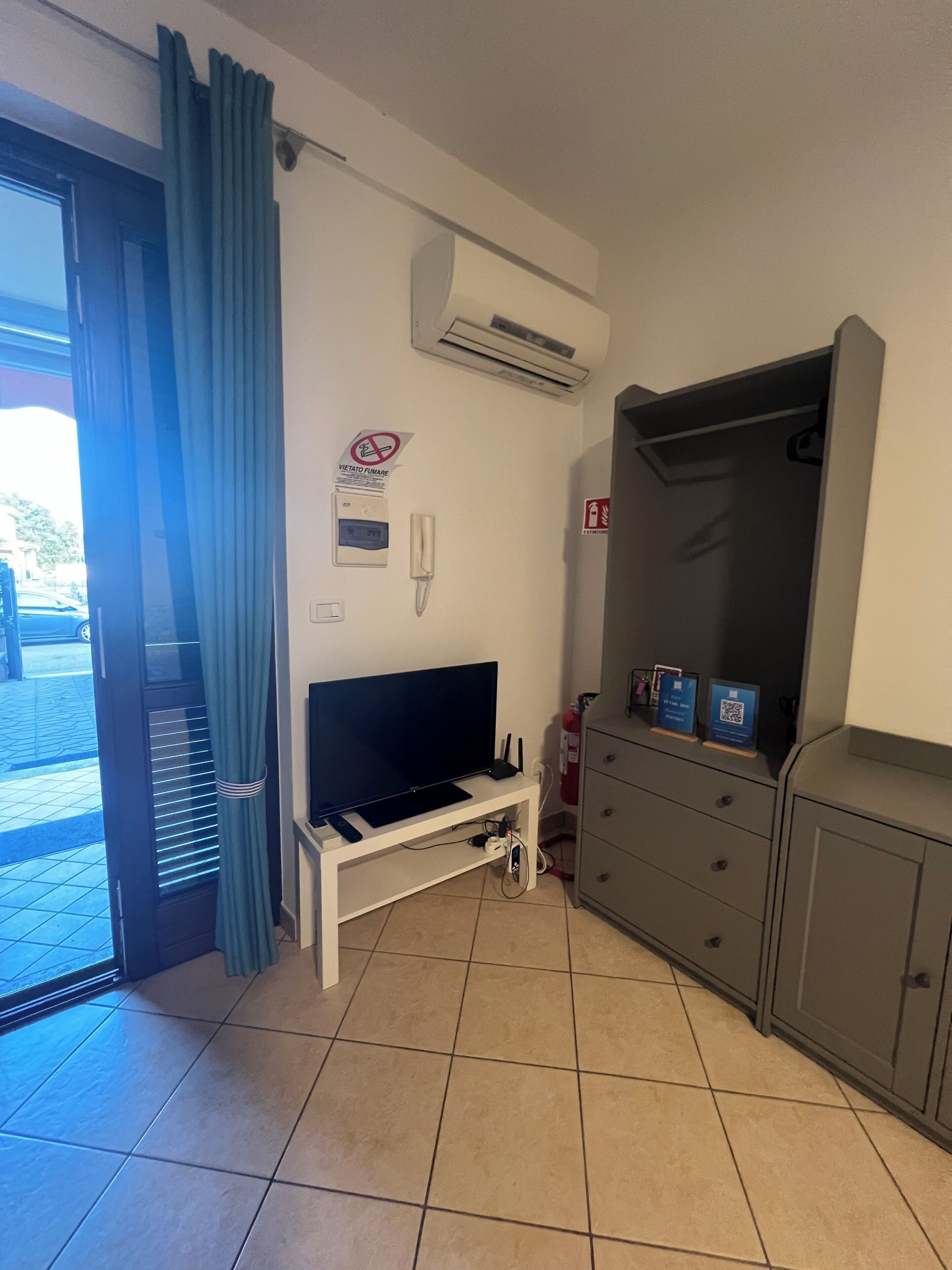 Quarto com televisão em um suporte, ar condicionado branco, extintor de incêndio e porta com cortina azul.