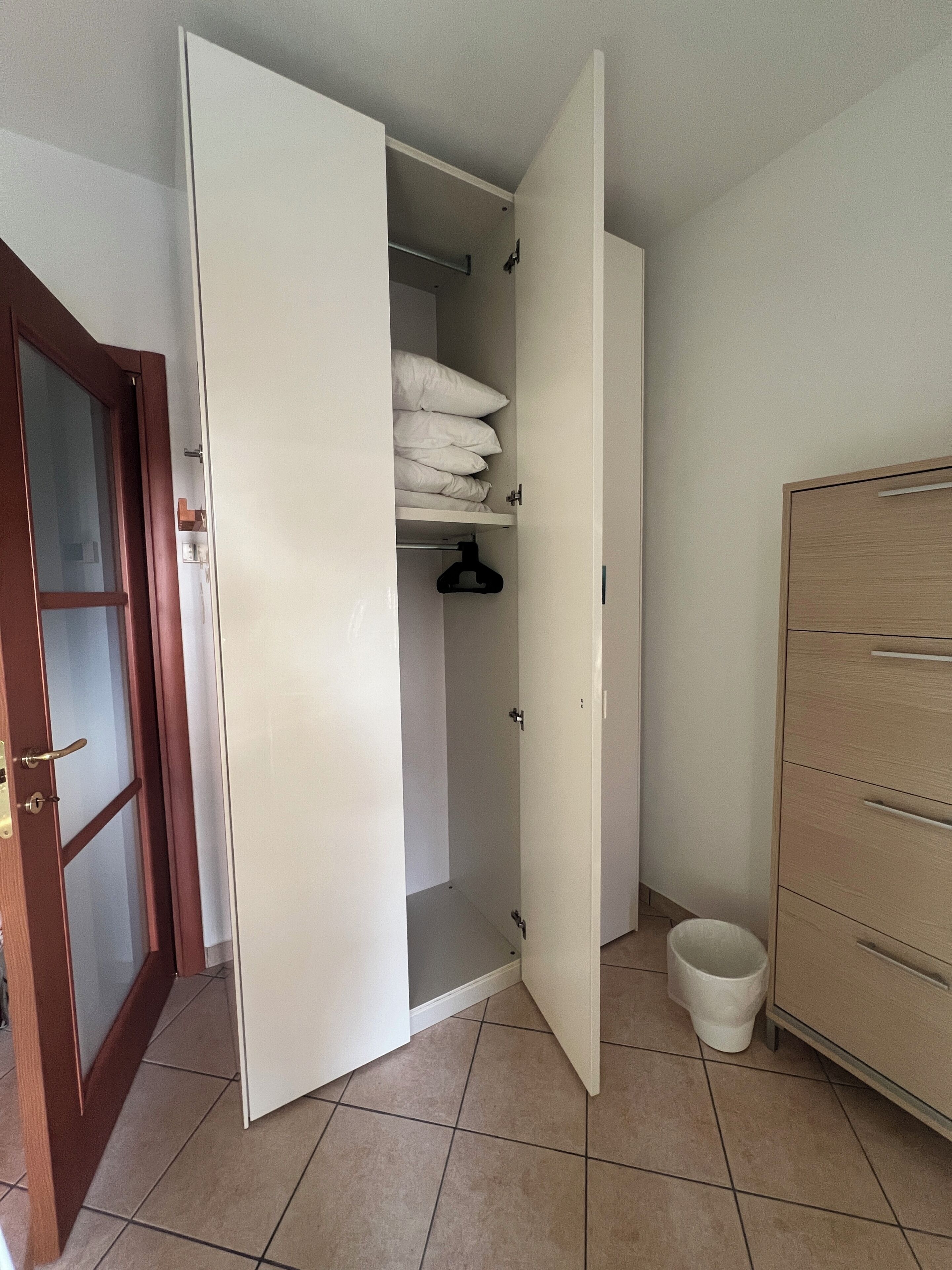Een kamer met een kledingkast, een klein plankje met kussens, een hangrek en een witte emmer.