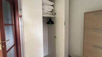 Een kamer met een kledingkast, een klein plankje met kussens, een hangrek en een witte emmer.