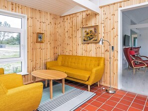Ein Zimmer mit einem gelben Sofa, einem kleinen runden Tisch und einem roten Sessel. Es gibt zwei Fenster und eine Tür zu einem anderen Raum.