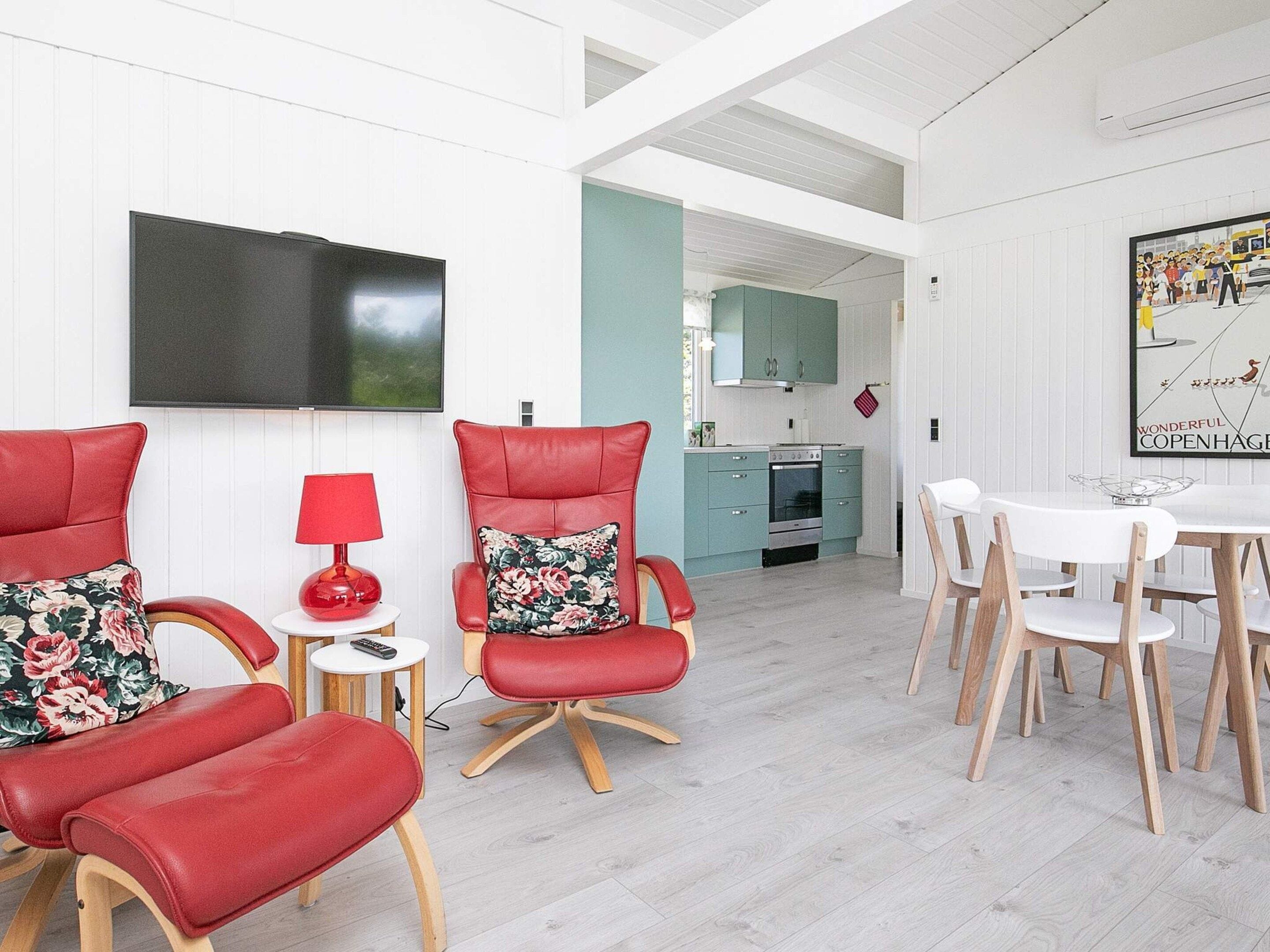 A modern living room with red leather chairs, a flat-screen TV, a dining table with white chairs, and a kitchen in the background.