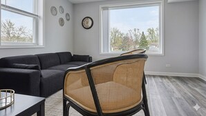 Una habitación con un sofá de color oscuro, una silla de mimbre, una mesa de comedor y un ventanal con vistas a árboles y edificios.