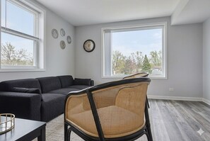 Ein Zimmer mit einem dunkelbraunen Sofa, einem Korbstuhl, einem Esstisch und einem großen Fenster mit Blick auf Bäume und Gebäude.
