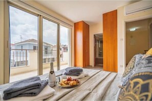 Ein Hotelzimmer mit großem Fenster, einem Bett mit gemustertem Überwurf, einem Nachttisch mit Flasche und Tablett mit Speisen sowie einer Wand mit Holzvertäfelung.