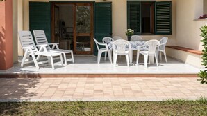An outdoor patio with white plastic chairs and a table, green shutters, and a tiled floor.