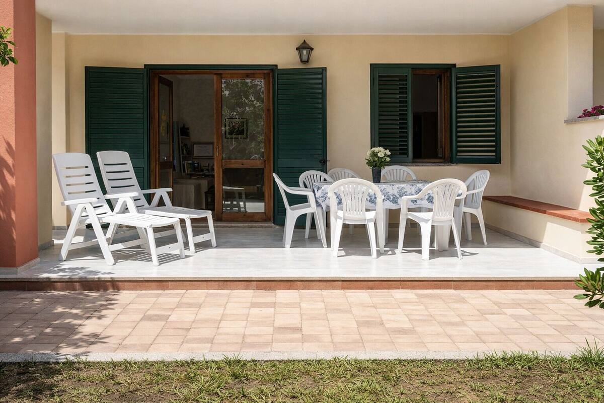 Una veranda esterna con sedie e tavolo in plastica bianca, persiane verdi e pavimento piastrellato.