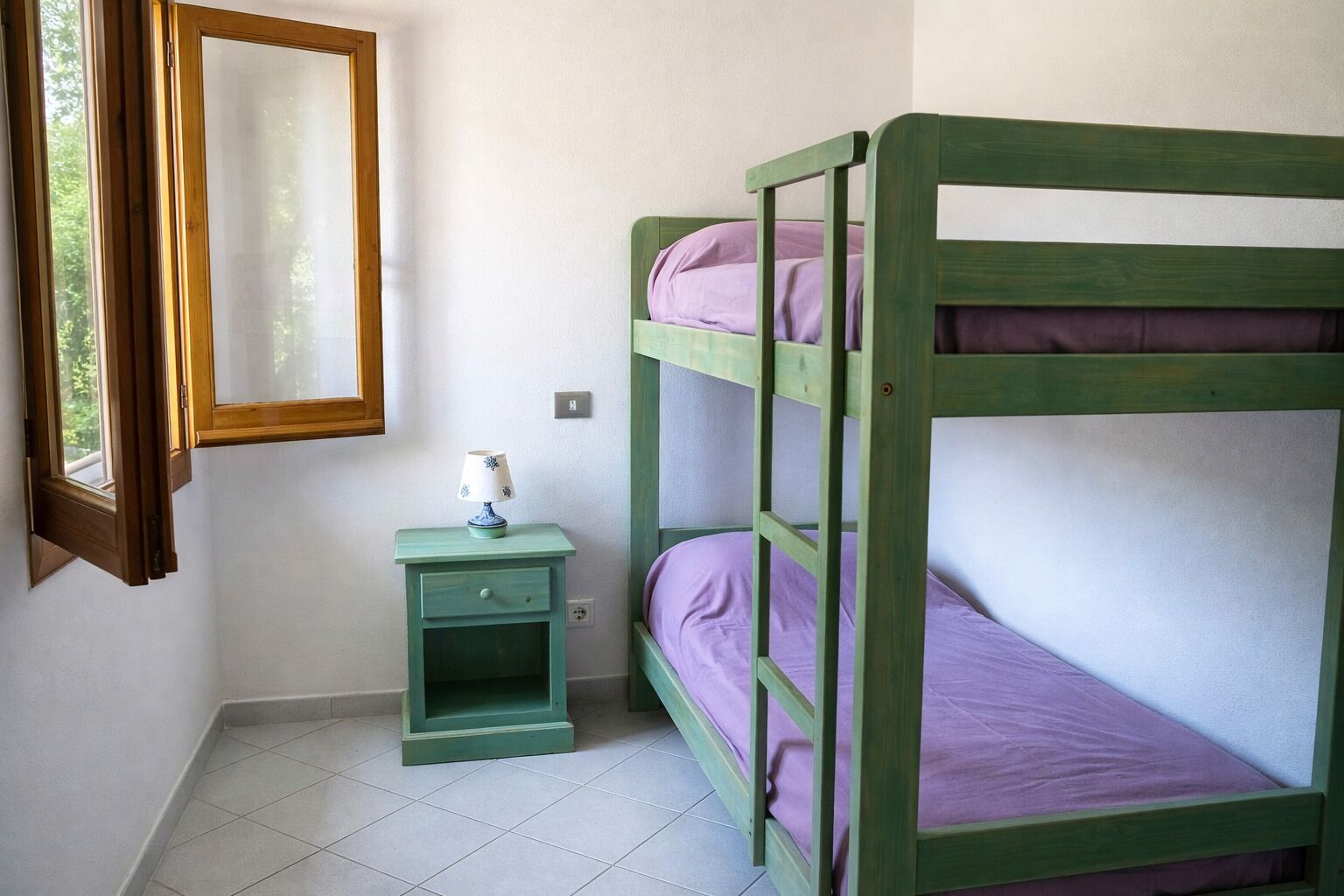 A bunk bed with a nightstand and a lamp, a window with a wooden frame, and a wall-mounted mirror.