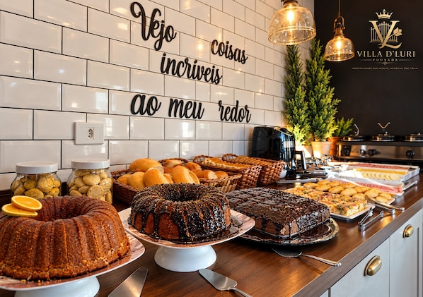 Un bufé con diversos productos de pastelería y postres, una cafetera y un cartel con un mensaje en portugués.