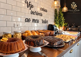 Buffet com diversos pães e doces, uma cafeteira e uma placa com mensagem em português.