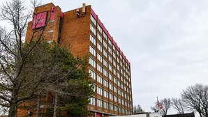 Um edifício de vários andares em tijolo, com telhado vermelho, entrada coberta e área de estacionamento com carros.