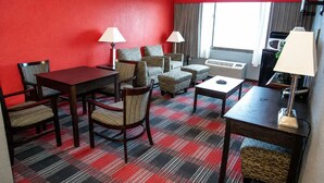 Quarto de hotel com parede vermelha, carpete xadrez, mesa de jantar com cadeiras, conjunto de sofá, duas luminárias e janela com cortinas.