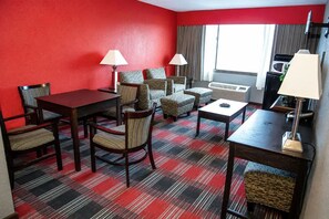 Hotellihuone, jossa on punainen seinä, ruutukuviollinen matto, ruokapöytä tuoleineen, sohvaryhmä, kaksi lamppua ja ikkuna, jossa on verhot.