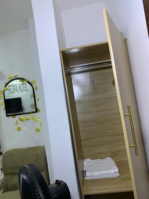 Quarto com guarda-roupa de madeira, cama com manta branca, sofá e espelho fixado na parede com a placa 'FEJPA HOTEL'.