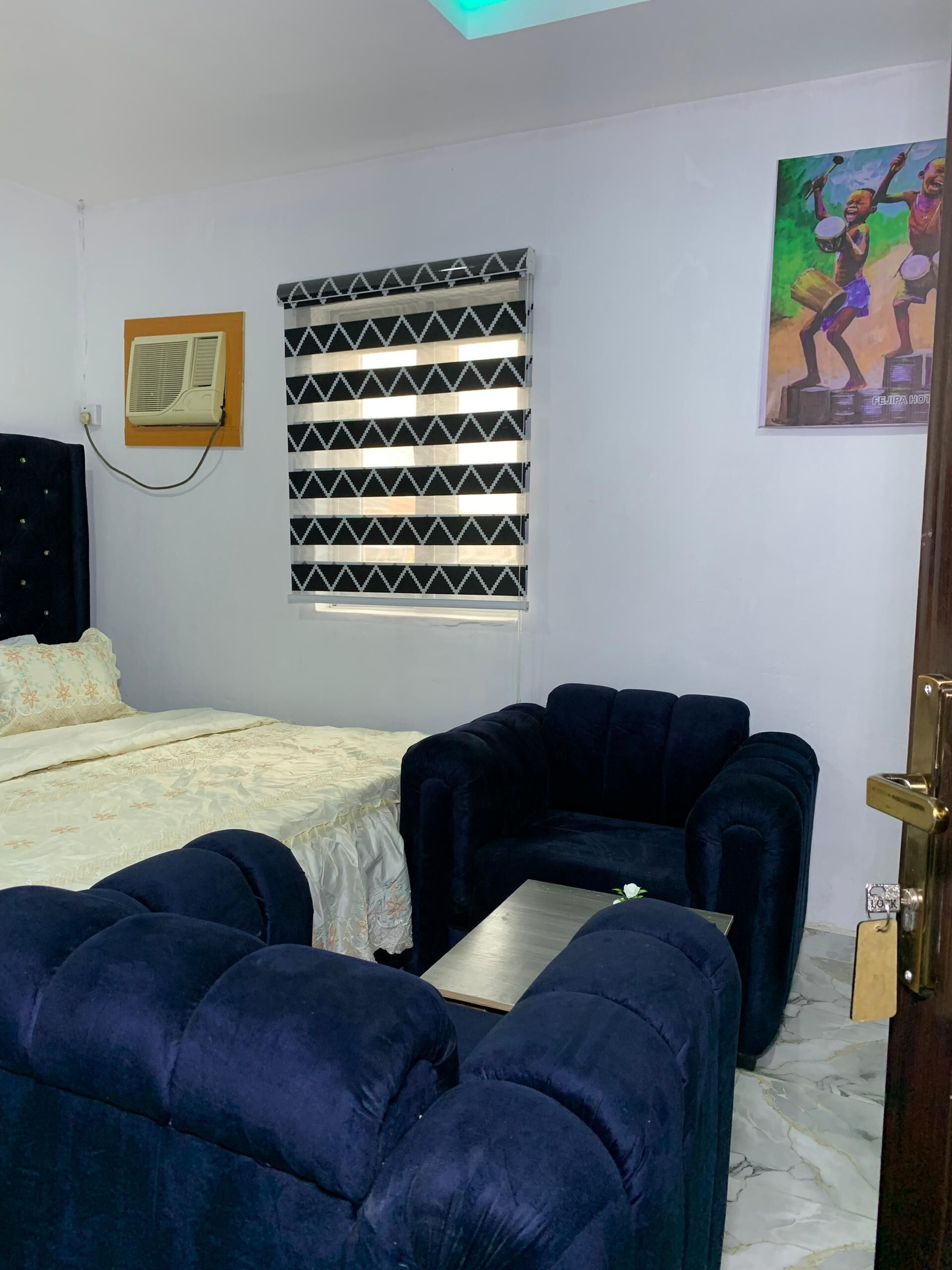 Een kamer met een bed, een bank, een kleine tafel, een raam met zonwering, een airconditioning en een schilderij aan de muur.