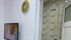 Um banheiro com chuveiro, pia, televisão fixada na parede e porta que dá para outro cômodo.